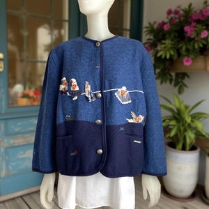 Könnte beinhalten: Blaue Wolljacke mit rundem Kragen und Knopfverschluss. Die Jacke hat ein zweifarbiges Design mit einem dunkleren blauen Boden und gestickten Details von Vögeln und Wäsche. Die Jacke wird auf einer Schaufensterpuppe präsentiert.