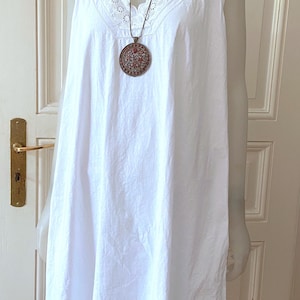May include: White sleeveless nightgown with a V-neckline and lace trim. A round pendant with a detailed design hangs from a brown cord. The nightgown is displayed on a mannequin.