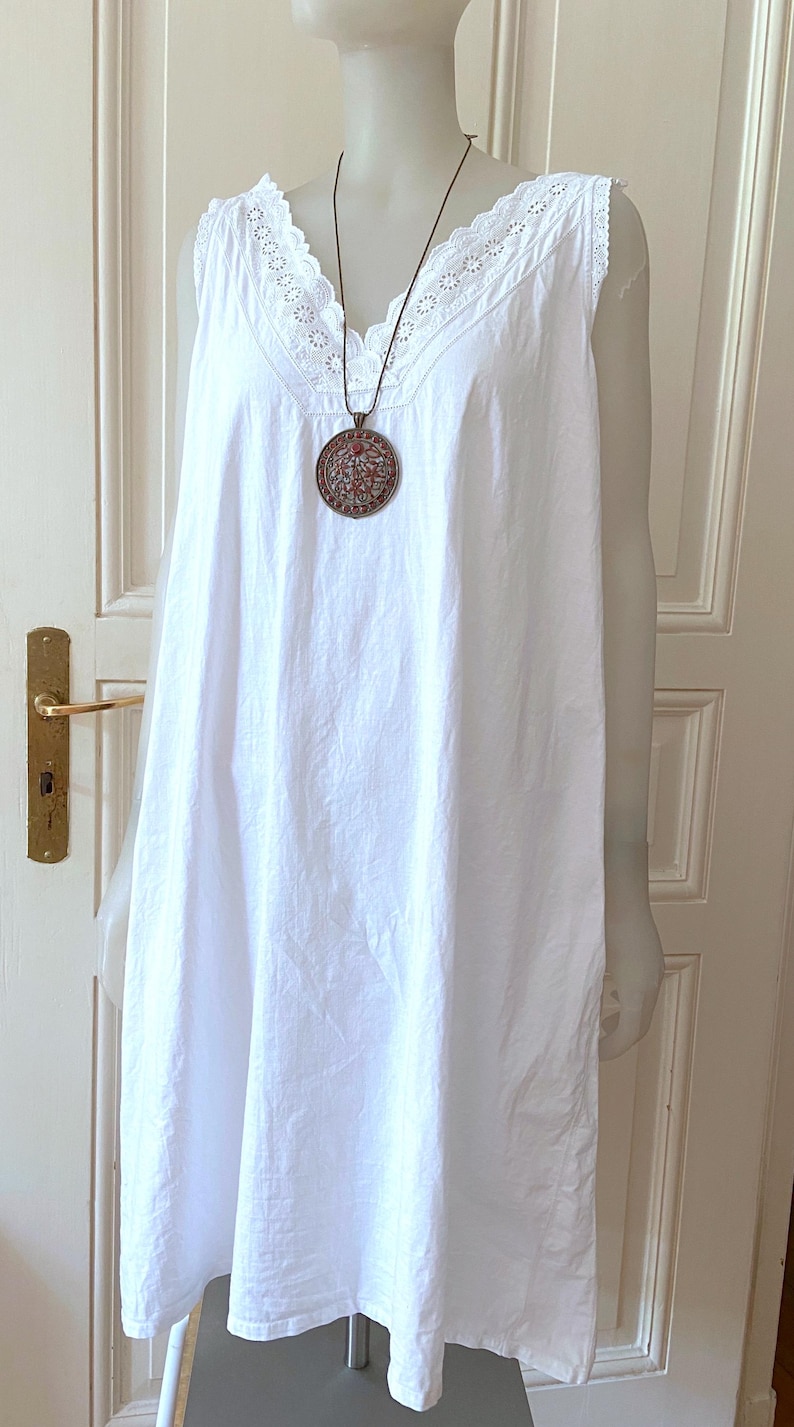 May include: White sleeveless nightgown with a V-neckline and lace trim. A round pendant with a detailed design hangs from a brown cord. The nightgown is displayed on a mannequin.