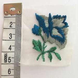 May include: A blue and green embroidered flower applique on a white fabric background. The flower is about 2 inches wide.