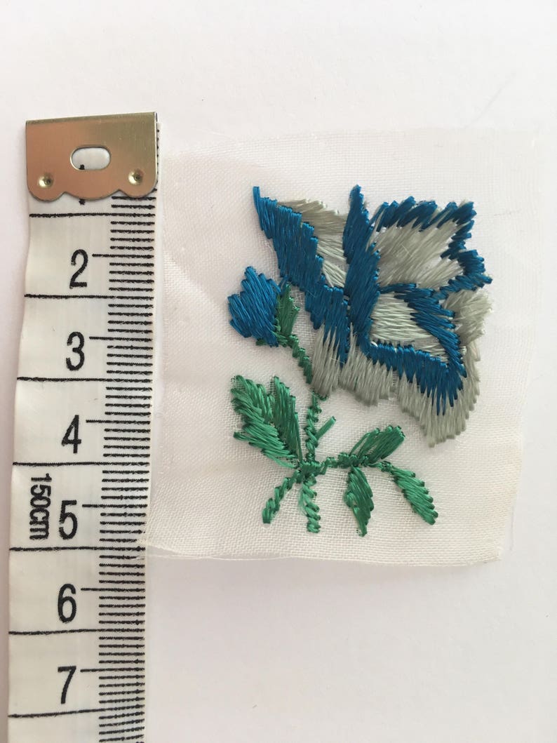 May include: A blue and green embroidered flower applique on a white fabric background. The flower is about 2 inches wide.