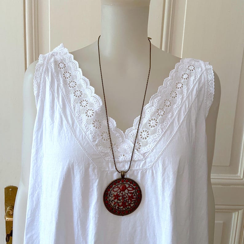 May include: A white sleeveless top with a V-neckline, adorned with delicate lace trim. A long, dark brown necklace with a circular pendant featuring a red floral design hangs from the neckline. The pendant has a bronze-colored frame.