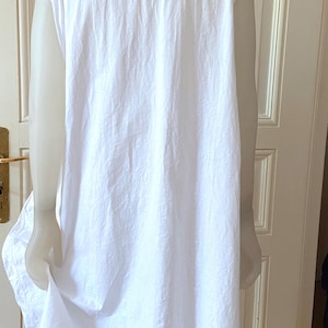 May include: A white, sleeveless nightgown with a scoop neckline and delicate lace trim. The garment is made of a lightweight fabric and hangs loosely. The nightgown is displayed on a mannequin, showcasing its simple, classic design.