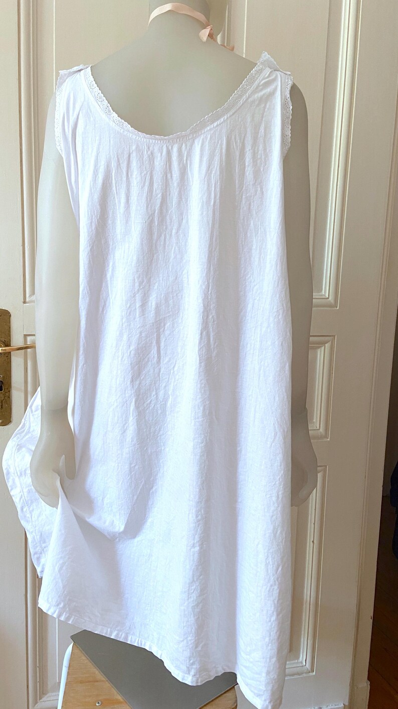 May include: A white, sleeveless nightgown with a scoop neckline and delicate lace trim. The garment is made of a lightweight fabric and hangs loosely. The nightgown is displayed on a mannequin, showcasing its simple, classic design.