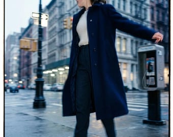 May include: A long navy blue coat worn over a cream-colored ribbed sweater and dark trousers, paired with brown heeled ankle boots. The outfit features a structured collar, button-down front, and a tailored fit, capturing a classic urban aesthetic.