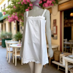 May include: A white cotton slip dress with lace trim along the neckline and hem. The dress has wide straps and a simple, classic design. The dress is displayed on a mannequin in an outdoor setting with a blurred background.