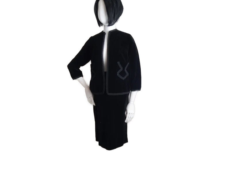 May include: A black velvet skirt suit with a cropped jacket featuring a contrasting black trim. The jacket has a V-neckline and long sleeves.