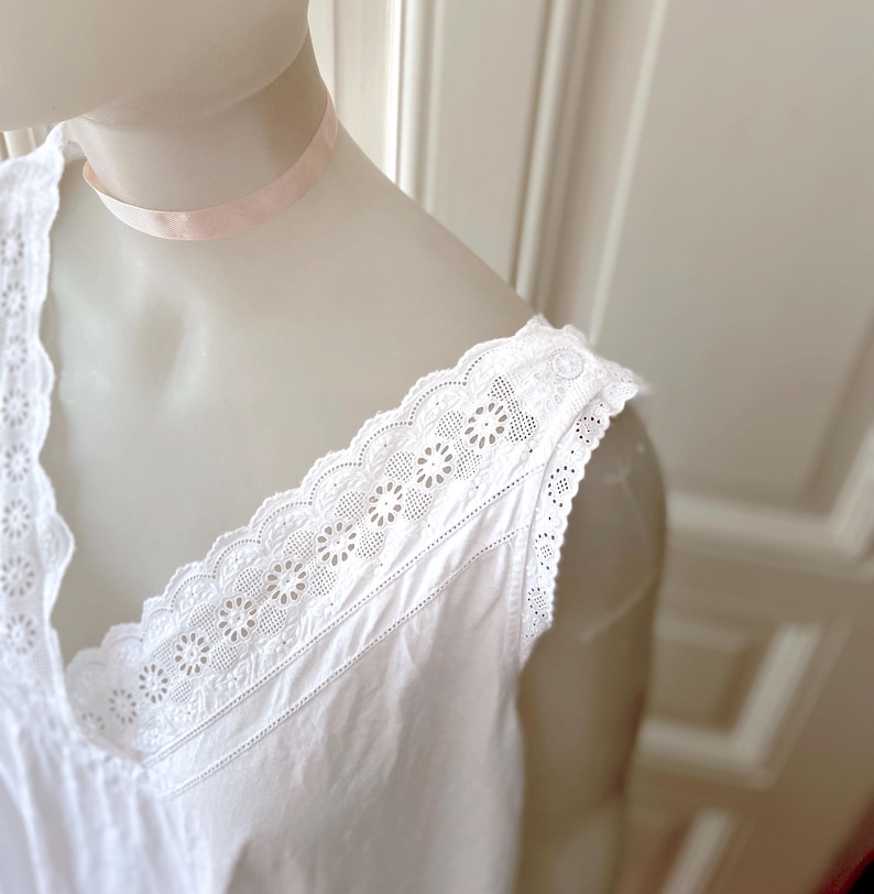 May include: White sleeveless nightgown with a V-neckline and delicate floral lace trim along the neckline and shoulders. The nightgown is displayed on a mannequin, showcasing its soft, flowing fabric and vintage design.
