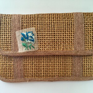 May include: A woven straw clutch with a brown trim and a blue and white embroidered flower.