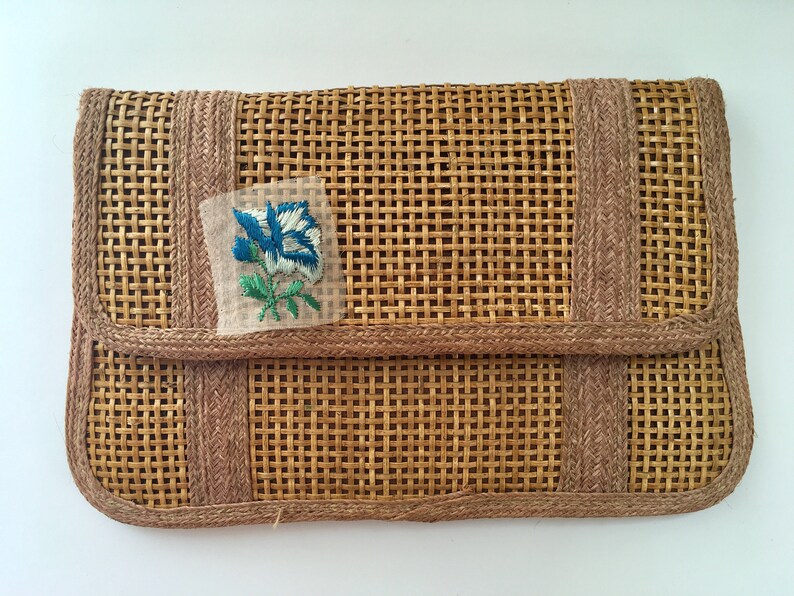 May include: A woven straw clutch with a brown trim and a blue and white embroidered flower.