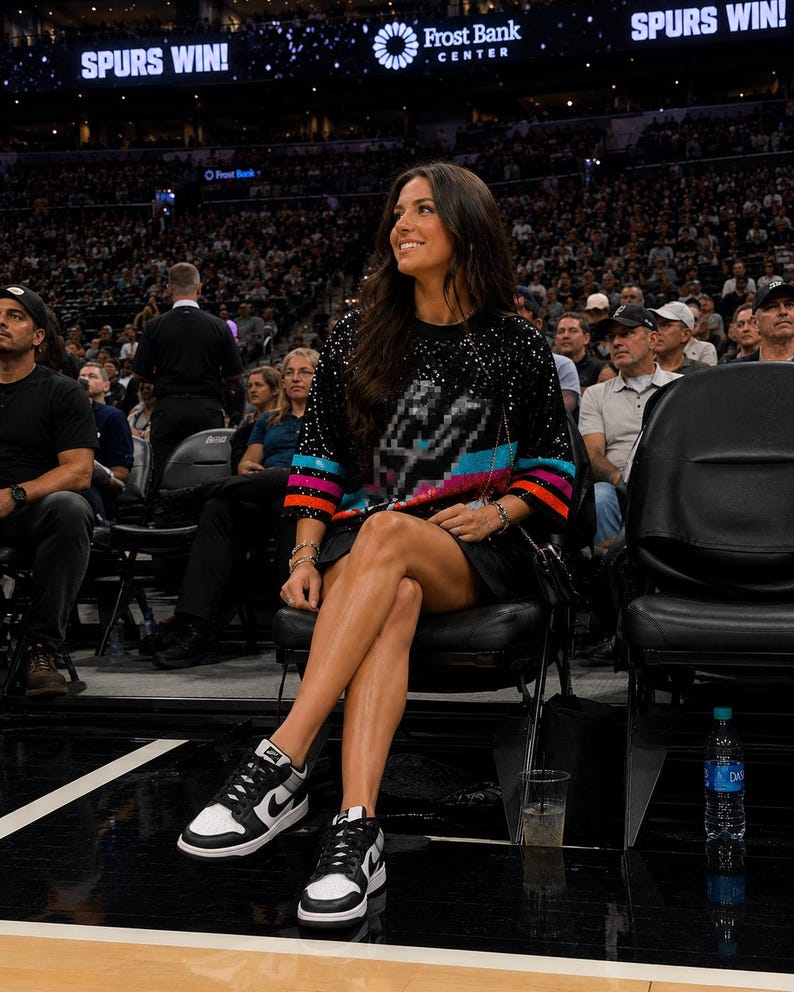 May include: A black long-sleeve sweater featuring a pixelated graphic of the San Antonio Spurs logo on the chest, accented with horizontal pink, blue, and orange stripes on the sleeves, paired with black sneakers with white panels and black laces.