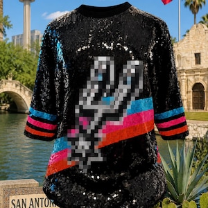 May include: A black sequined t-shirt featuring a pixelated silver spur logo on the front. The sleeves and lower torso are accented with horizontal stripes in bright blue, pink, and orange. The garment has a round neckline and short sleeves.