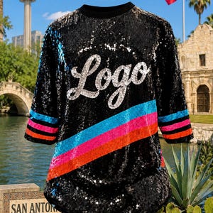May include: A black sequin shirt featuring the word Logo in silver script across the chest. The garment includes a diagonal stripe pattern in bright blue, pink, and orange across the front and on the sleeves, with a solid black crew neckline.