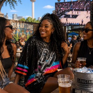 May include: A black sequined t-shirt featuring a pixelated San Antonio Spurs logo on the front and horizontal stripes in blue, pink, and orange on the sleeves. The shirt is worn in an outdoor setting with a basketball game visible on a screen.