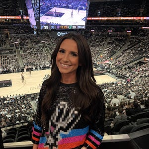 May include: A black long-sleeved shirt covered in sequins featuring a pixelated silver spur logo on the chest with horizontal stripes of blue, pink, and orange across the midsection, worn in a large indoor sports arena with a scoreboard overhead.