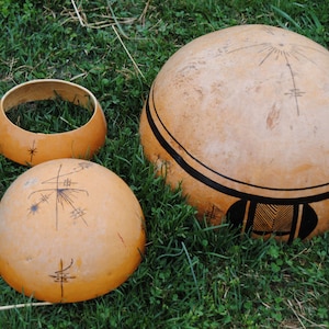 May include: Three gourds with intricate black and white designs. The largest gourd is round and has a black band around the middle. The other two gourds are smaller and have a more bowl-like shape. All three gourds are sitting on a bed of green grass.