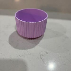May include: A light purple, cylindrical container with vertical ribbed detailing. The interior is a slightly darker shade of purple. The container sits on a light-colored surface. The object appears to be made of a flexible material.