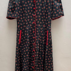 May include: A black midi dress featuring a repeating pattern of small red roses and green leaves. The dress has short sleeves, a V-neckline, a front button closure with red buttons, and two vertical side pockets with red trim along the openings.