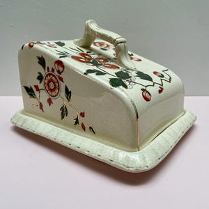 Puede incluir: Fuente de queso de cerámica color crema con tapa y asa. El plato está decorado con motivos florales rojos y verdes y detalles dorados. La base es rectangular con un borde festoneado.