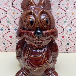 May include: A brown glazed ceramic biscuit jar shaped like a rabbit wearing overalls. The rabbit has large eyes, pointed ears, and a black nose. The word biscuits is embossed on the front of the overalls in raised, rounded lettering.
