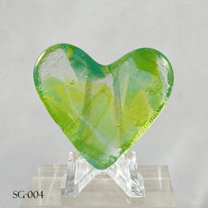 May include: A heart-shaped glass ornament featuring a marbled pattern of lime green, emerald, and translucent white colors with visible air bubbles throughout the material. The object is displayed on a clear acrylic stand with the text SG-004.