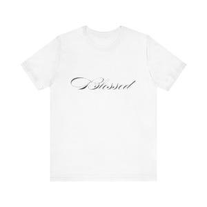 以下が含まれることがあります： 胸に筆記体で「Blessed」と書かれた白いTシャツ。テキストは濃いグレーです。シャツは半袖でクルーネックです。
