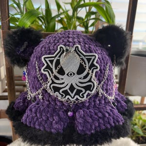 May include: A purple crocheted hat with black faux fur trim and ear-like accents. The hat features a silver octopus emblem with chain and bead embellishments. The hat is a unique accessory.