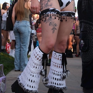 May include: White crochet leg warmers featuring black tie closures with fuzzy pom-poms and a black faux fur trim at the bottom, paired with black platform boots, a black and white cow print bodysuit, and a black lace garter with star charms.