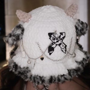May include: A white crocheted bucket hat featuring two light beige horns on top, a black and white cow-print X-shaped patch, and a silver-toned chain embellishment with small charms. The brim is trimmed with a fluffy black and white faux fur.