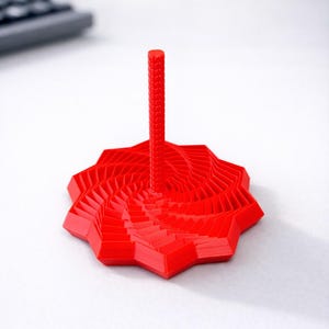 以下が含まれることがあります： 鮮やかな赤色の3Dプリントされた卓上玩具。星型のベースにスパイラル模様、中央にテクスチャード加工された柱が特徴です。白い背景に置かれ、モダンな美学を暗示しています。