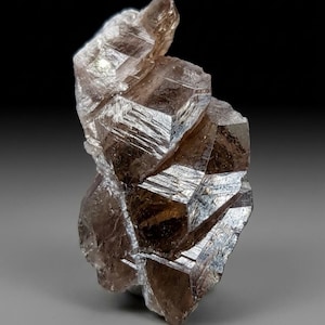 Puede incluir: Primer plano de una formación cristalina marrón con una estructura compleja y en capas. El cristal tiene una apariencia facetada, con múltiples planos que reflejan la luz. La forma general es irregular, con una parte superior puntiaguda y una superficie rugosa y texturizada.