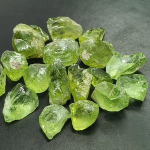 Puede incluir: Una colección de piedras preciosas de peridoto en bruto y sin cortar. Las piedras son de un color verde vibrante y translúcido, con formas irregulares y texturas rugosas. Las piedras preciosas están dispersas sobre una superficie oscura.