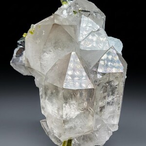 Puede incluir: Un grupo de cristales de cuarzo transparente con puntas geométricas y afiladas. Los cristales tienen una cualidad translúcida, con toques de verde claro e inclusiones negras. La estructura general es compleja y multifacética, mostrando formaciones geológicas naturales.