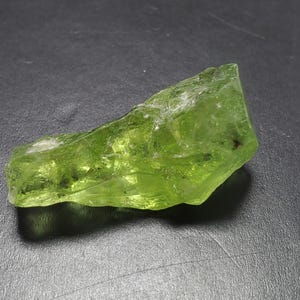 Puede incluir: Una gema de peridoto en bruto, translúcida, con un tono verde claro vibrante. La piedra en bruto tiene una estructura cristalina y una forma irregular, con algunas inclusiones oscuras. La gema se muestra sobre un fondo oscuro.