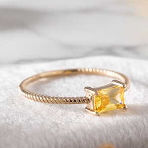 May include: A gold ring with a yellow rectangular gemstone. The ring has a twisted band and a square setting for the gemstone. The background is a soft white surface with decorative elements.
