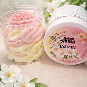 Peut inclure: Un pot transparent rempli de crème fouettée rose et jaune en couches, ressemblant à une fleur. À côté, un récipient cylindrique avec une étiquette portant le texte "Maison Giulia JASMIN" et des illustrations de fleurs de jasmin. Des fleurs de jasmin blanches au premier plan.
