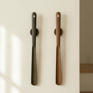 Puede incluir: Dos calzadores de madera, uno marrón oscuro y otro marrón claro, montados en una pared. Cada calzador tiene un mango largo y delgado con un pequeño agujero en la parte superior y un extremo redondeado para facilitar la inserción del zapato. Los calzadores están fijados a la pared con soportes circulares de color marrón oscuro.