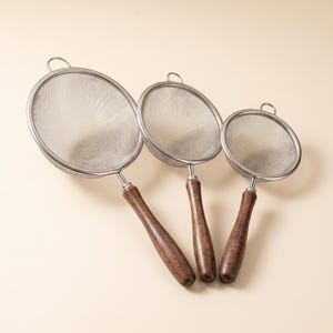 May include: Three stainless steel mesh strainers with wooden handles. The strainers are of varying sizes, with a circular mesh head and a loop at the top for hanging. The wooden handles are a dark brown color.