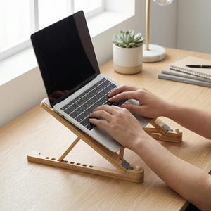 May include: A laptop computer is positioned on a natural bamboo laptop stand. The stand is angled for ergonomic use. The laptop is open, and the user's hands are on the keyboard. The stand is designed to elevate the laptop for better viewing.