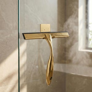 May include: A gold-colored shower squeegee with a black rubber blade is mounted on a glass shower door. The squeegee has a curved handle and a square mounting bracket. The background shows a steamy shower environment.