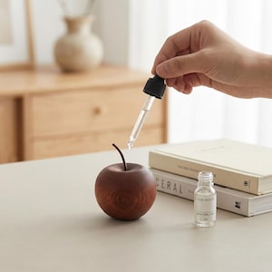 Peut inclure: Une pomme en bois avec une tige est posée sur une table. Une pipette en verre transparent est tenue au-dessus de la pomme, avec une goutte de liquide qui tombe. Une petite bouteille en verre avec le texte "JAM MOON" et "POOL ONE DAY" est à côté de la pomme.