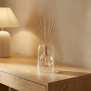 Puede incluir: Un difusor de vidrio transparente con varillas de madera. El difusor tiene una base redondeada y una cubierta transparente en forma de cúpula. Las varillas están dispuestas en el centro del difusor. Una lámpara color crema es visible en el fondo, sobre una superficie de madera.