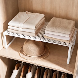 May include: A white wire shelf unit with folded clothing and a straw hat. The shelf holds stacks of beige and tan sweaters and shirts. A straw hat with a brown band sits below the shelf. The items are inside a wooden closet.