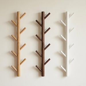 May include: Three wooden coat racks in a row, each with a tree-like design. The racks are in natural, dark brown, and white finishes. Each rack has multiple pegs extending outwards, ideal for hanging coats or accessories. The racks are mounted on a neutral wall.