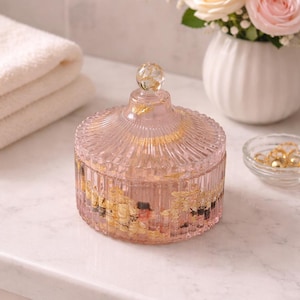 May include: A pink glass jar with a lid, adorned with gold leaf accents. The jar has a ribbed design and a clear glass knob on top. The jar is sitting on a white surface, with a vase of roses in the background.