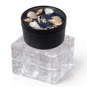 May include: A black round container filled with blue wax and seashells, resting on a clear glass block. The seashells vary in size, shape, and color, adding a natural aesthetic. The container is a decorative item.