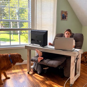 May include: A white wooden desk on wheels with a laptop and a black monitor placed on top. The desk is positioned in front of a brown recliner chair. A golden retriever stands on the hardwood floor to the left of the desk, while another dog rests on the floor to the right.