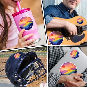 K&ouml;nnte beinhalten: Eine Sammlung runder Aufkleber mit einem farbenfrohen Berglandschaftsdesign mit roter Sonne, Regenbogenbergen und dunkelblauen B&auml;umen. Die Aufkleber sind auf einem rosa Becher, einer Gitarre, einem Baseballhelm und einem Laptop abgebildet.