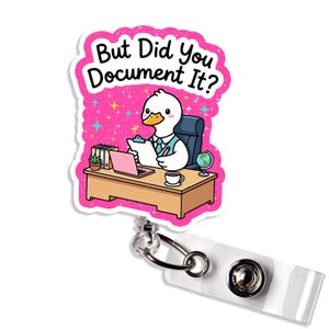 May include: A pink badge reel with a cartoon duck at a desk, holding a clipboard. The text "But Did You Document It?" is above the duck. The reel has a white strap and a metal clip.