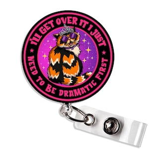 May include: A retractable badge reel with a cartoon cat wearing sunglasses and a tiara. The cat is orange, black, and white, set against a purple and pink background with stars. The text reads "I'll get over it I just need to be dramatic first."